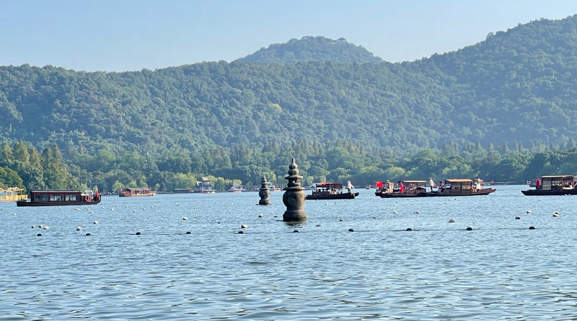 图片展示一座宁静的湖泊，湖中央有石塔，背景是群山和天空，湖面上散布着游船。