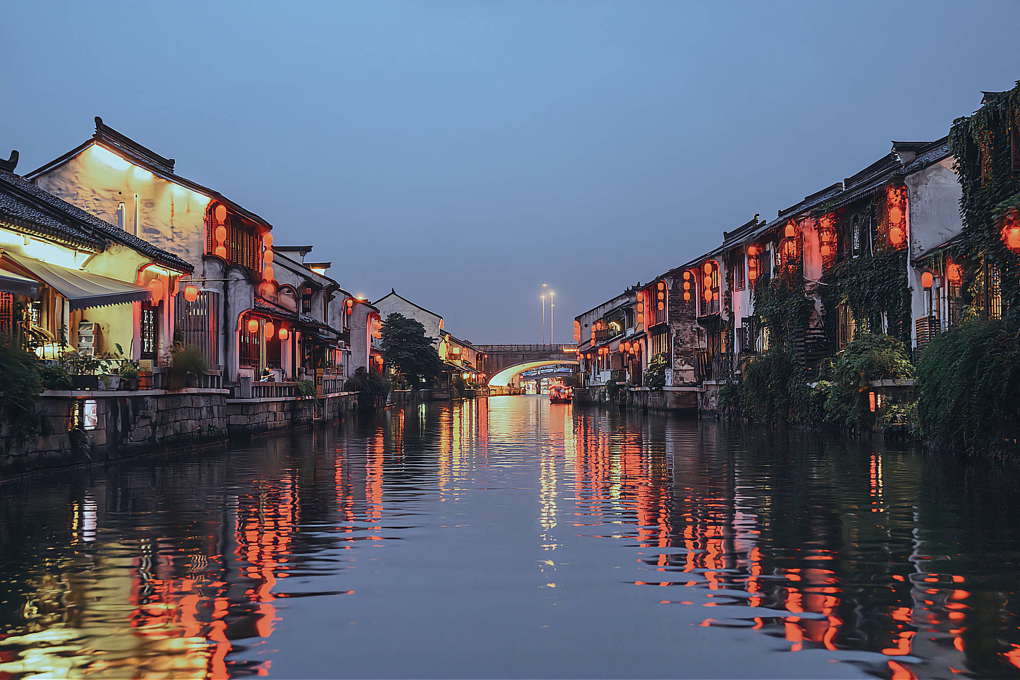 这是一幅古镇夜景图，古建筑沿河而建，灯光倒映在平静的水面上，远处有一座拱桥，营造出宁静和谐的氛围。