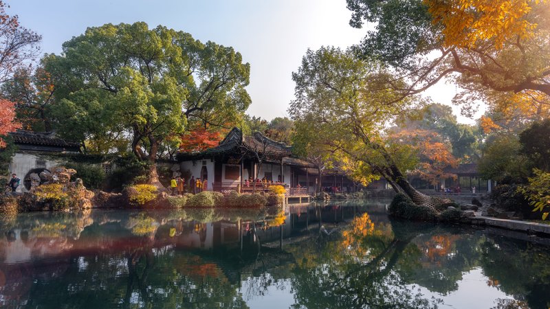 图片展示了一个宁静的中国园林，秋天的树木，古色古香的亭子，以及平静的湖面反射着周围的美景。