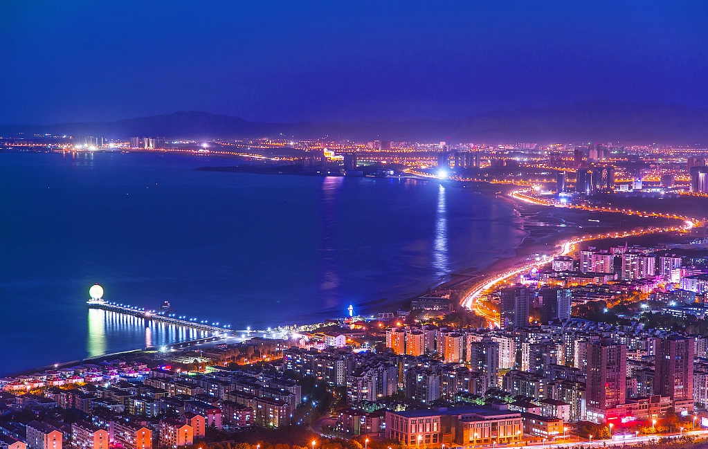 这是一张城市夜景照片，显示了灯火辉煌的建筑群、海滨、道路和一座照亮的摩天轮。天空呈现深蓝色调，远处可见山脉轮廓。