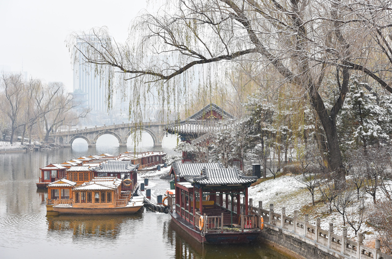 图片展示了雪后的中国古典园林，有拱桥、亭台和游船，远处轻雾笼罩，树木挂霜，呈现出宁静优美的冬日景色。