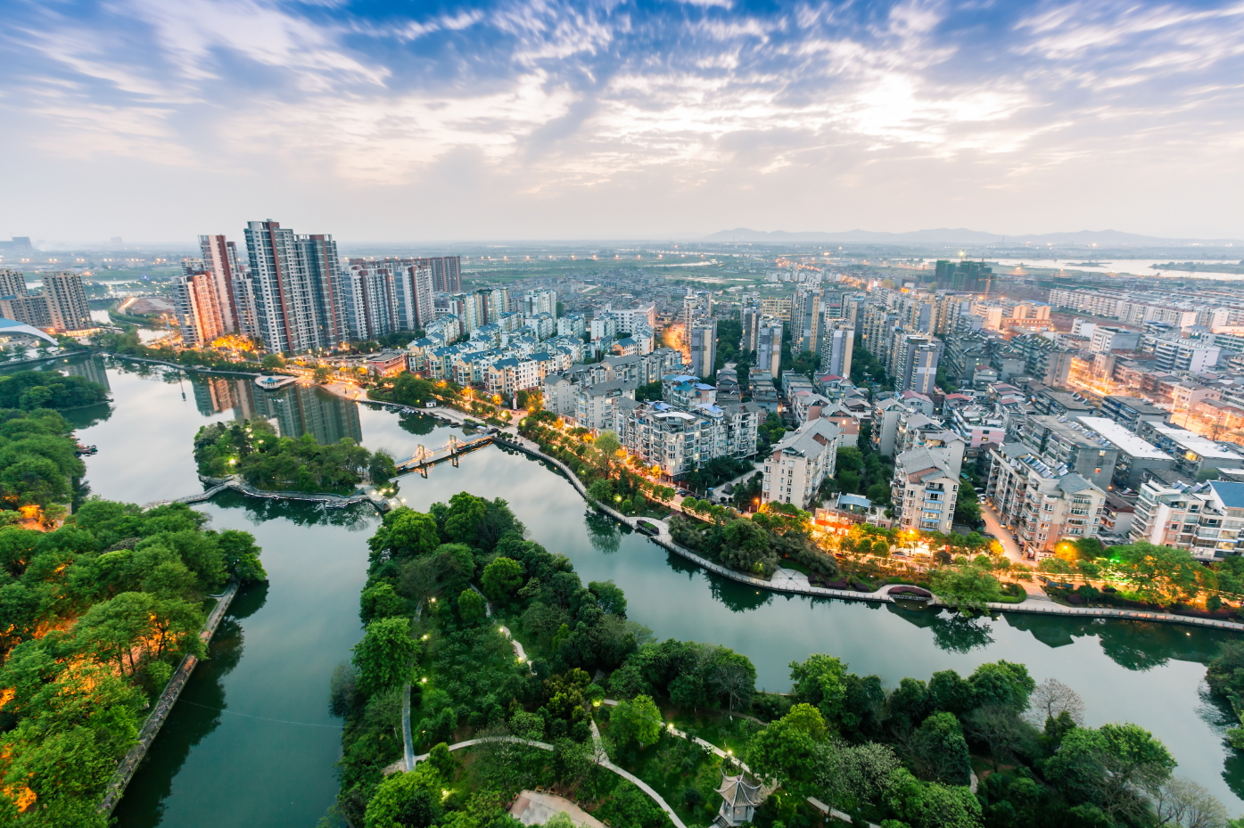 图片展示了一座现代城市的高空景观，有高楼大厦、绿树环绕的公园和蜿蜒的河流，天空呈现出日落的渐变色彩。