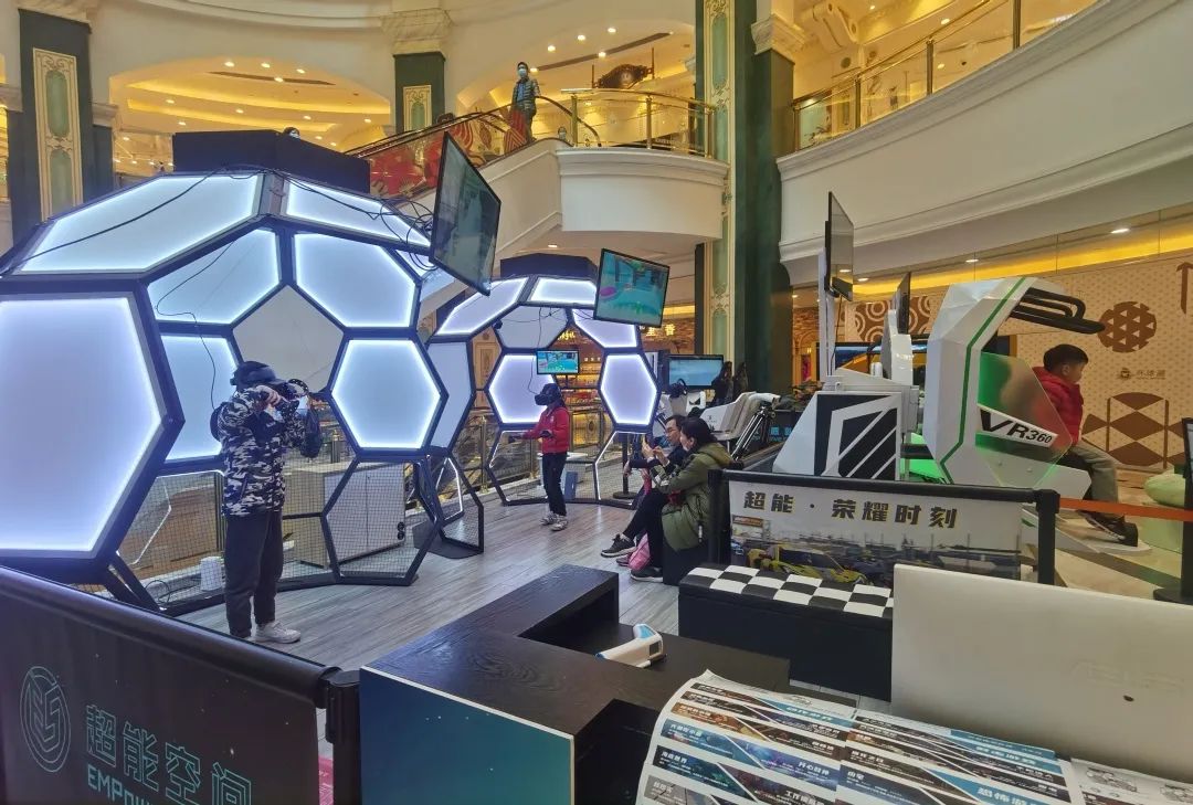 图片展示了一个商场内的电子游戏体验区，有多个六边形装饰的展台和几位顾客正在游玩或等待体验。