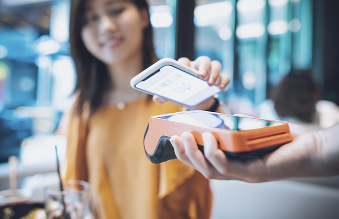 图片展示一位女士在使用手机扫码支付，她面前有一台橙色的移动支付设备，背景模糊，可能在一家商店或餐厅内。