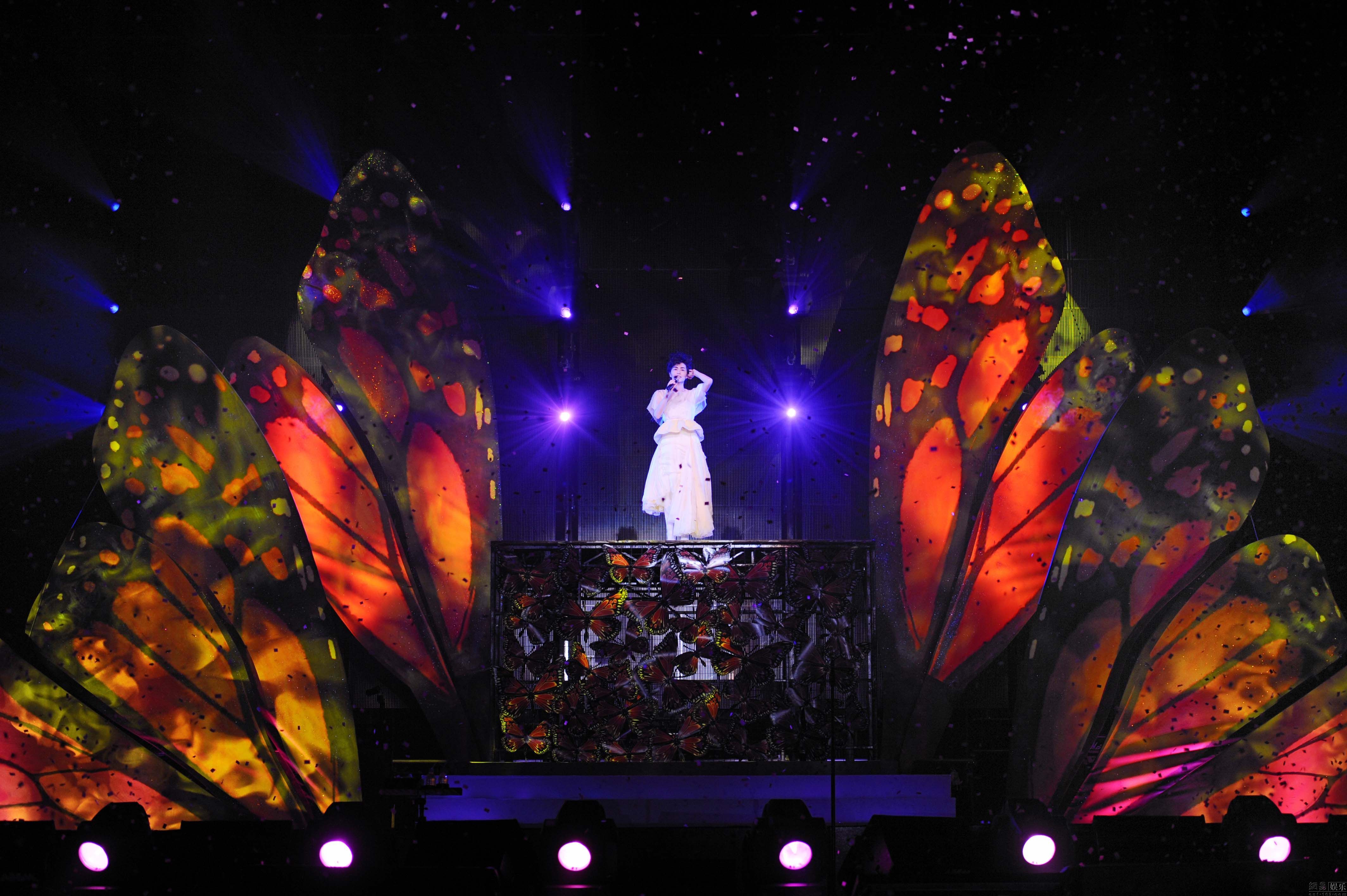 图片展示一位穿白色服装的人站在舞台中央，背后是巨大的彩色蝴蝶翅膀装置，灯光照耀下，气氛梦幻。