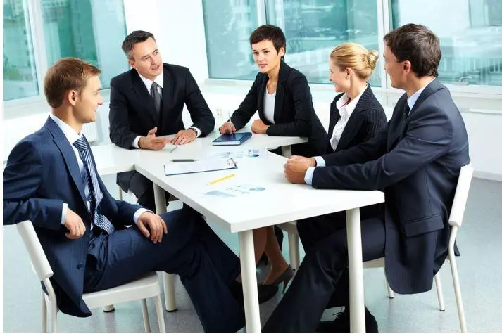 图片展示了五位穿着正装的职场人士正在办公室内围坐在会议桌旁，看起来正进行商务讨论或会议。