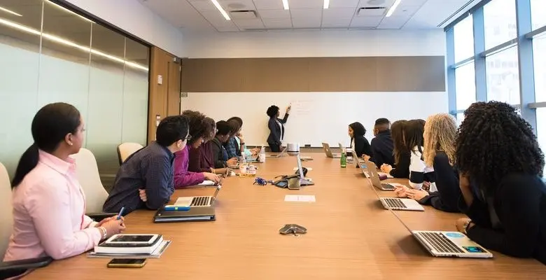图片展示了一间会议室，里面坐满了专注的听众，一位站立的人正在白板上做演示。环境正式，显得很专业。