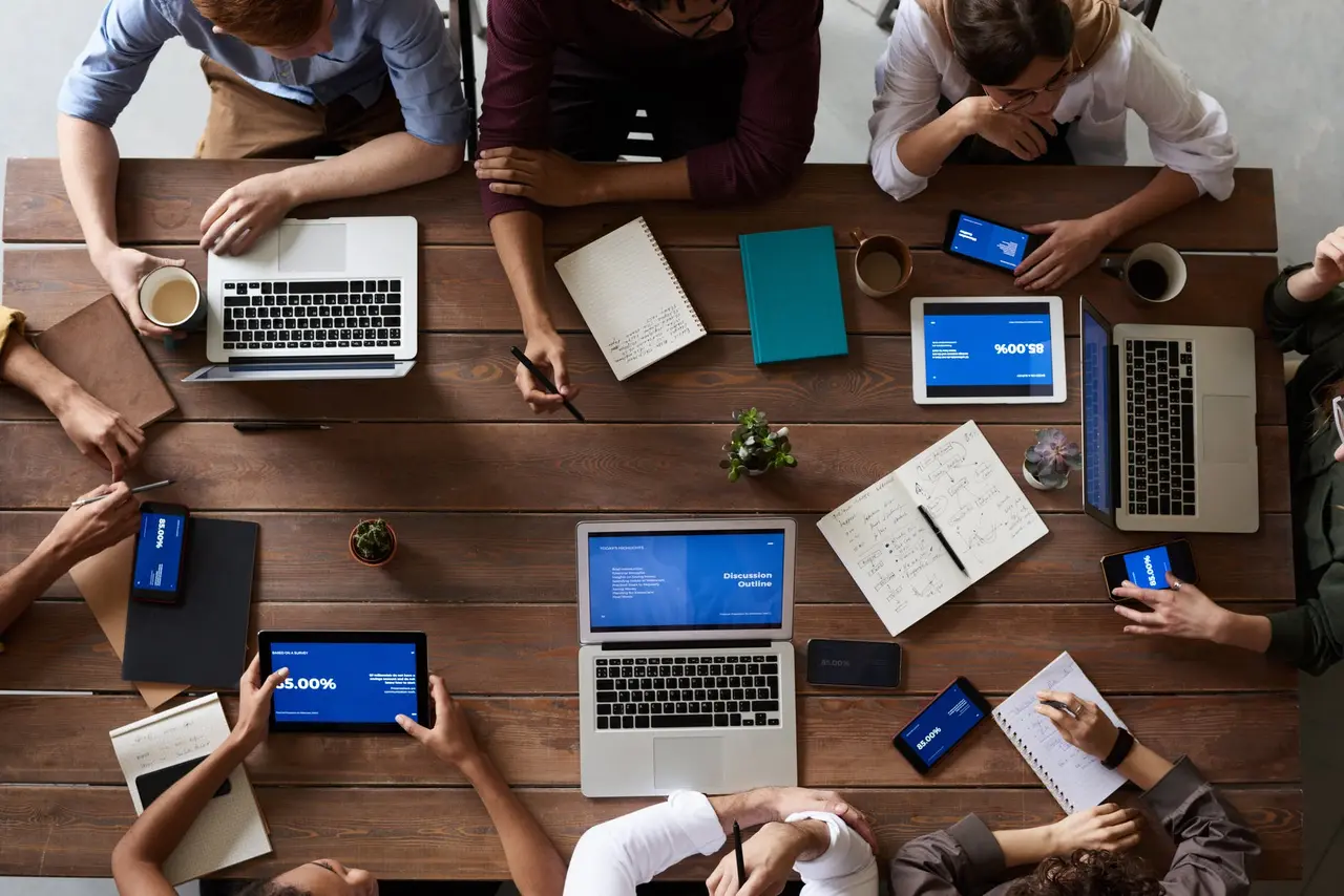 图片展示了多人围坐在会议桌旁，使用笔记本电脑和平板等设备，看起来正在进行商务会议或团队工作讨论。