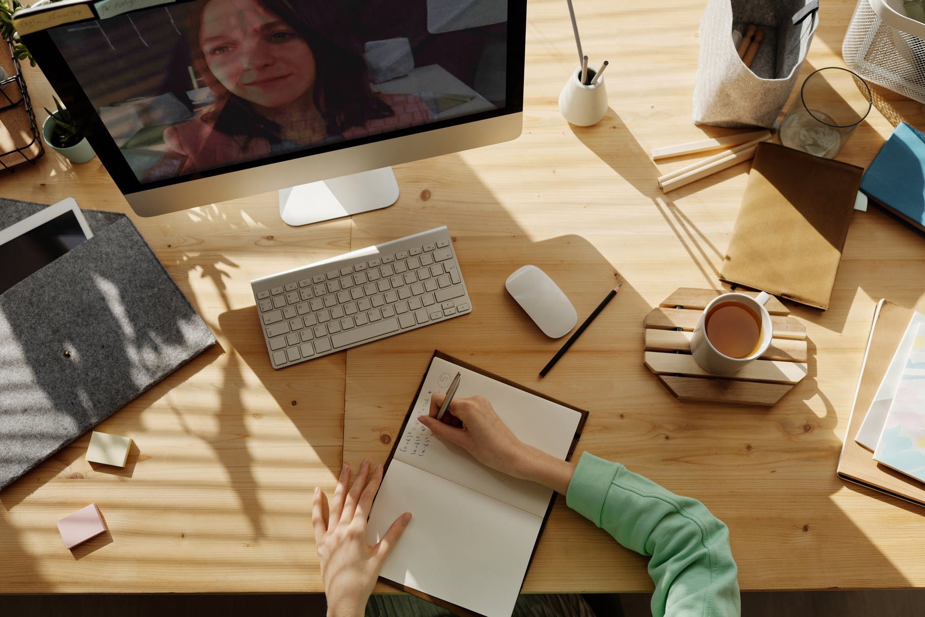 图片展示一位正在书桌前工作的人，电脑屏幕上有另一位女士的图像，桌面上散落着办公用品和一杯咖啡。