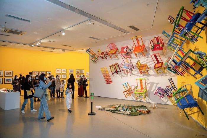 这是一间艺术展览厅，墙上挂有多彩椅子装置艺术，参观者在观看作品，展厅背景墙面呈黄色，氛围现代活跃。