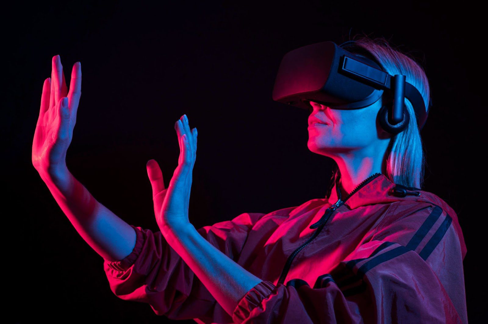 图片展示一位女性戴着虚拟现实头盔和耳机，似乎在体验VR内容，她举起双手，背景是暗色调，光线呈现出霓虹般效果。