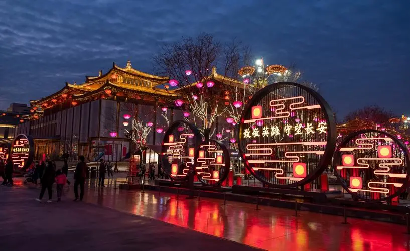 图片展示夜幕下的节日氛围，彩灯装饰，传统建筑背景，人们在欣赏，巨大圆形灯饰上有中国特色的图案和文字。