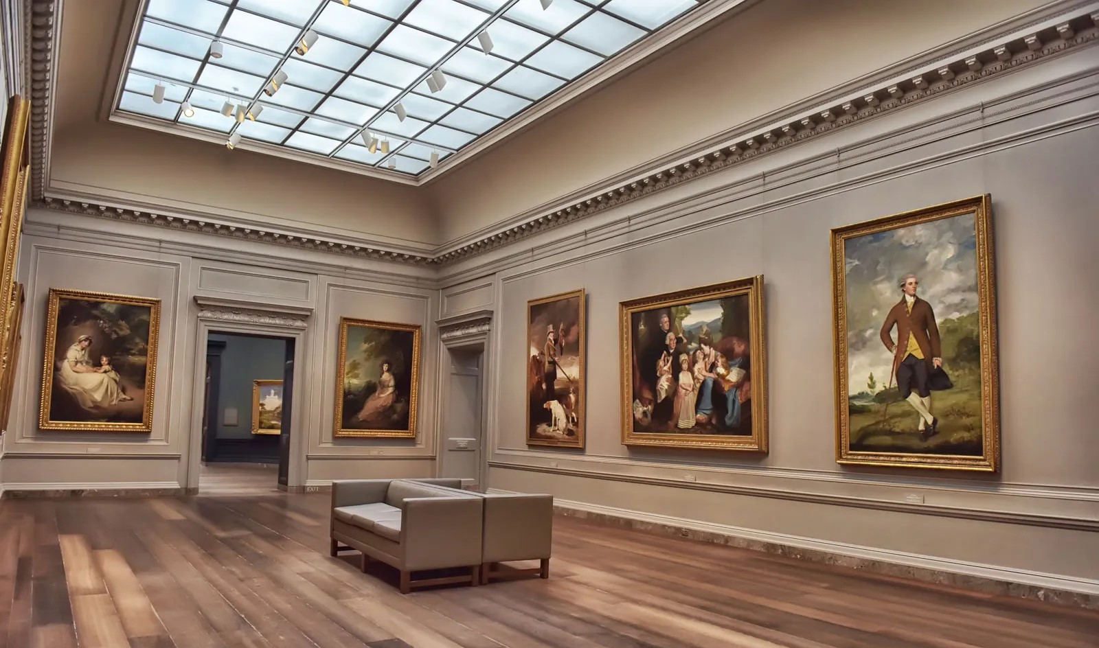 这是一间艺术画廊内部，墙上挂着不同风格的绘画作品，房间装饰典雅，有长椅和木质地板，顶部是透明天窗。