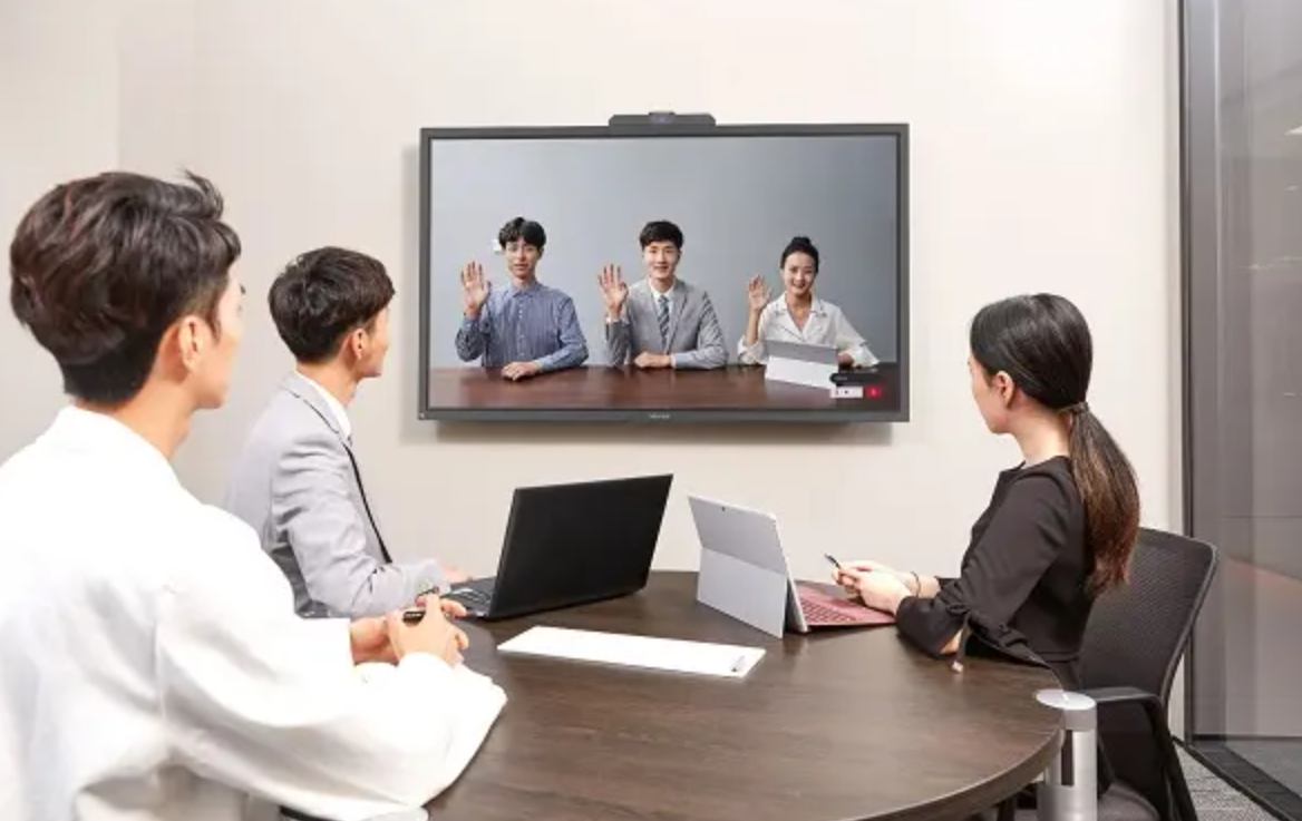 会议室内，四人围坐桌旁，前方大屏幕显示三人视频会议，其中一人挥手致意，现场氛围正式而友好。