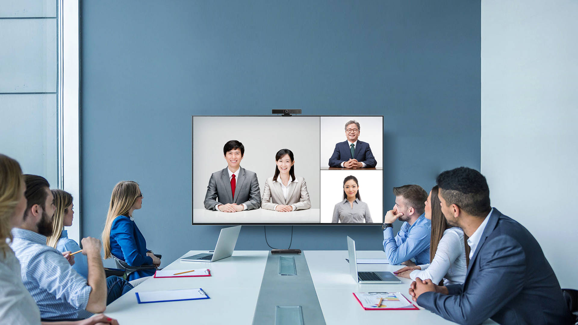 图片展示了一场视频会议，会议室内坐着几位专注的参与者，屏幕上显示着两位亚洲男女和一位老年男性。