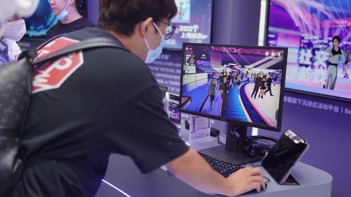 图片展示一位戴眼镜的男子正坐在电脑前，屏幕上显示着人群聚集的场景，背景是紫色调的科技感展览环境。
