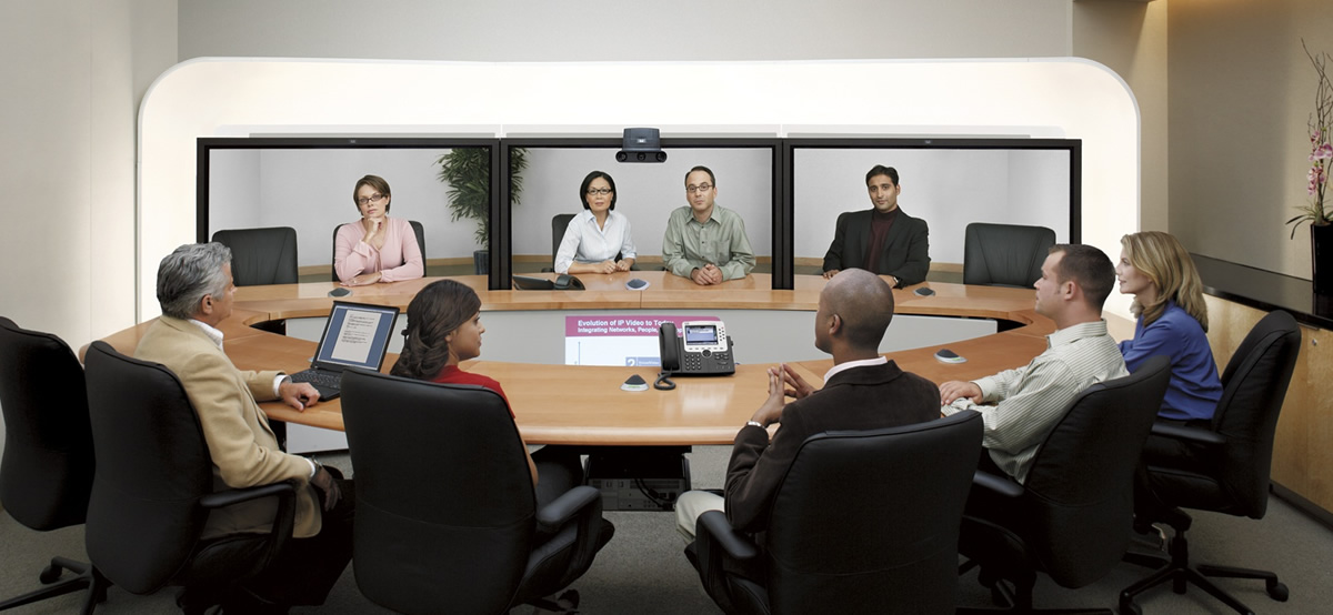 图片展示了一场视频会议，参与者坐在会议室的桌子两侧，屏幕中显示远程参与者的实时图像。