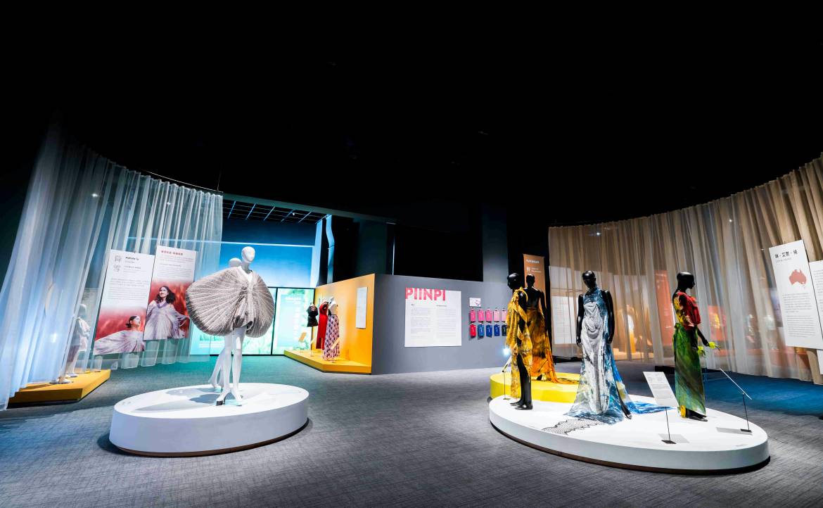 图片展示了一个时尚展览，有几个模特展示不同风格的服装，展厅设计现代，色彩鲜明，灯光聚焦在服装上。