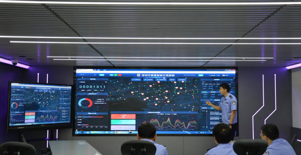 图片展示多人在高科技监控室内，一人站立汇报，其余坐听，墙上多个大屏幕显示数据和地图。