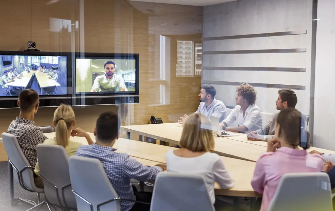 图片展示了一间会议室，多名参会者正坐着聚焦观看屏幕上的视频通话，通话中显示一位男士正在讲话。