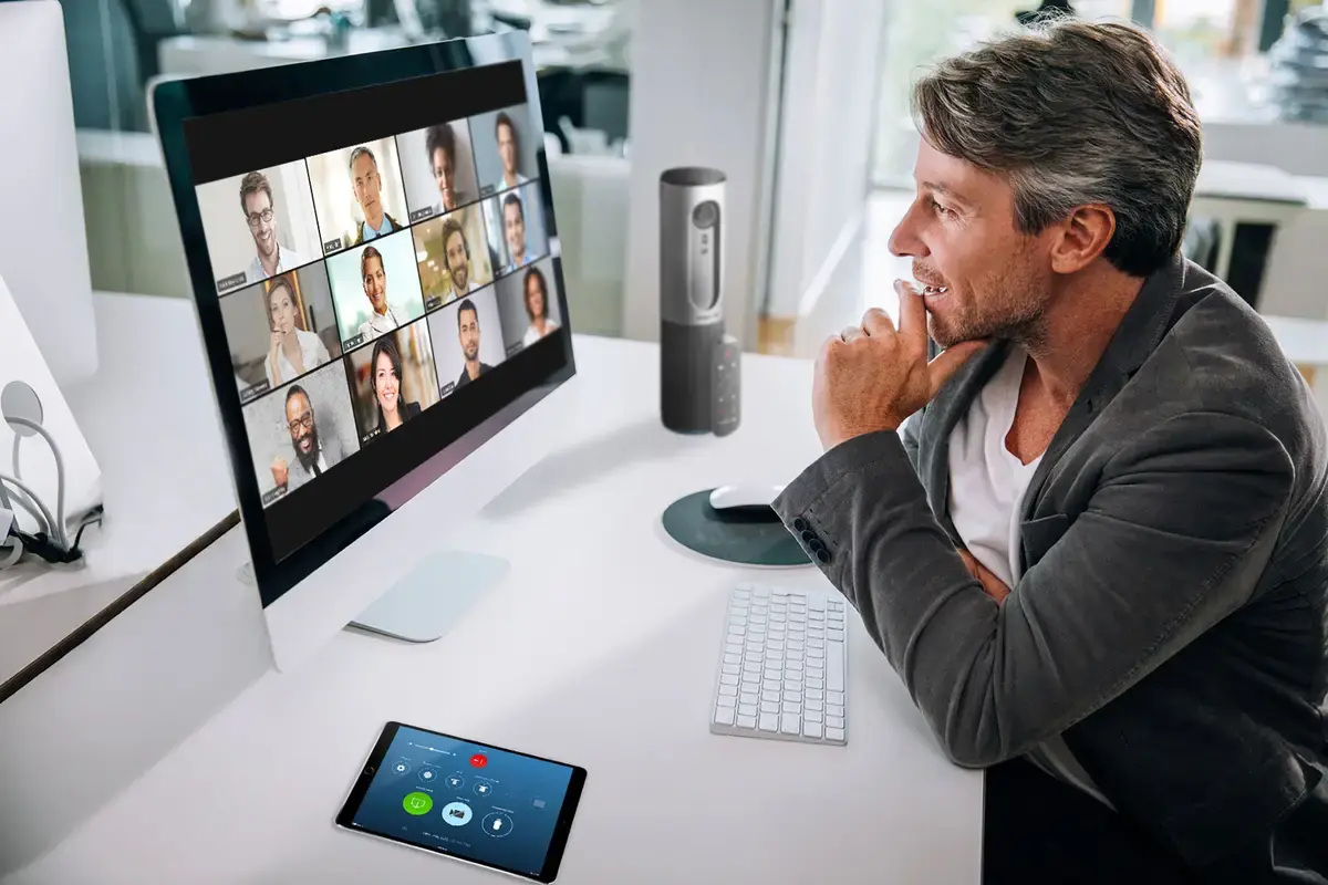 一位男士正专注地在电脑屏幕上观看多个视频会议窗口，似乎在进行远程工作或虚拟会议。办公环境现代，设备先进。