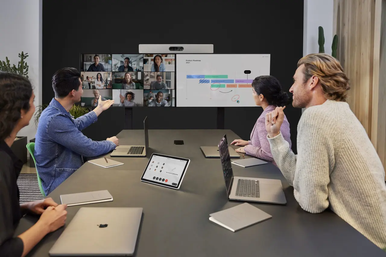 图片展示了四位专业人士在办公室进行视频会议，他们正通过大屏幕与远程参与者交流讨论工作内容。