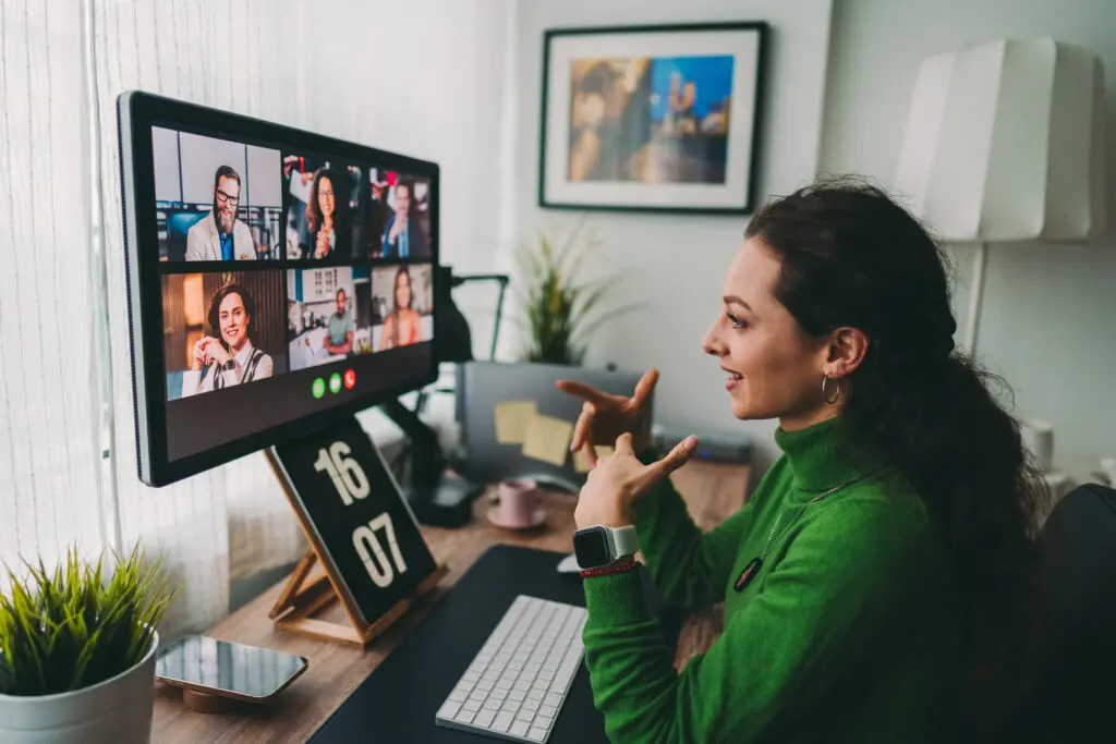 图片展示一位女士在家中使用电脑进行视频会议，屏幕上显示多位参与者，环境整洁，氛围专业。
