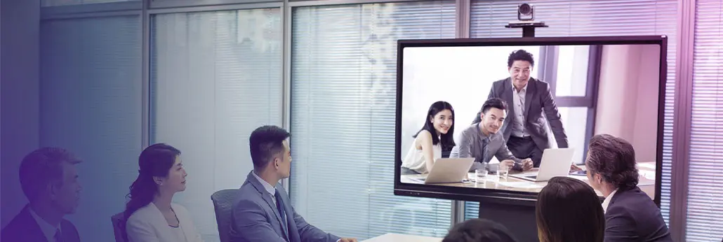 图片展示了一间办公室，几位职员正坐着观看大屏幕上的视频会议，屏幕中显示三位正在讨论的同事。
