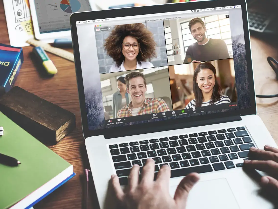 图片展示了一台笔记本电脑屏幕，屏幕上是一个视频会议界面，显示四位微笑的参与者，旁边是一些办公用品。