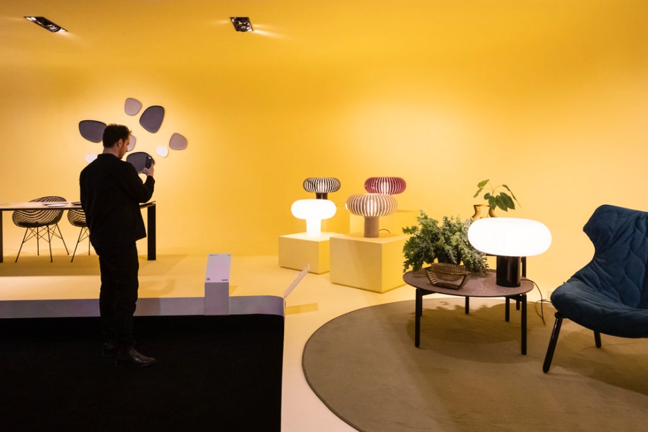 一位男士站在黄色背景的展览室内，观看墙上的艺术品。室内陈设有现代风格的家具和装饰植物。