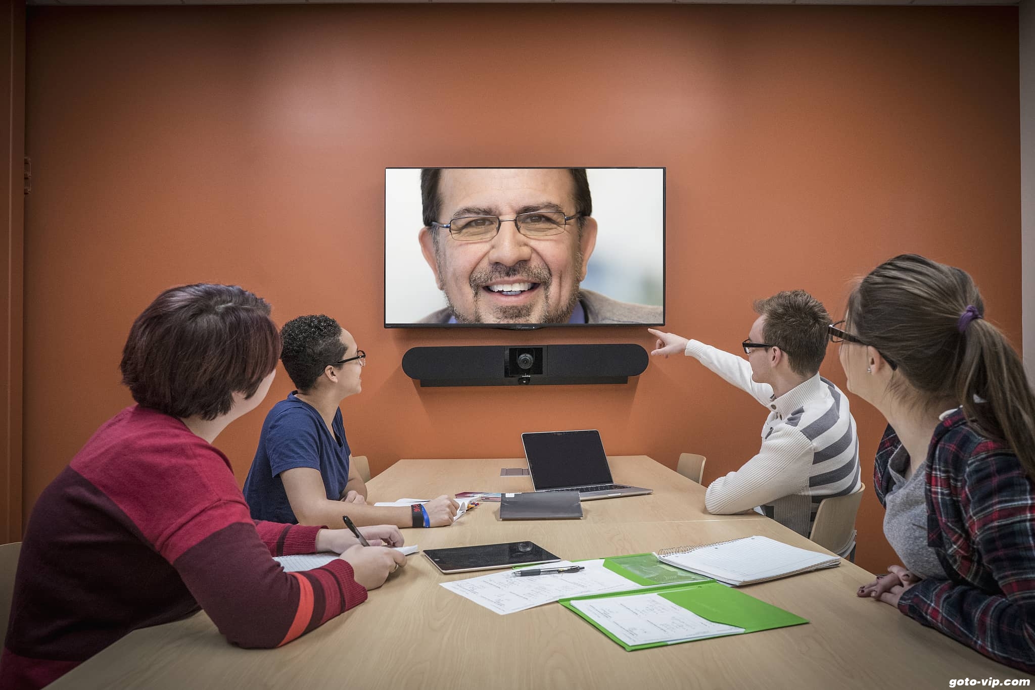 四人坐在会议室桌旁，正通过大屏幕视频通话，与远程的一位笑容可掬的男士交流。环境看起来正式且专业。