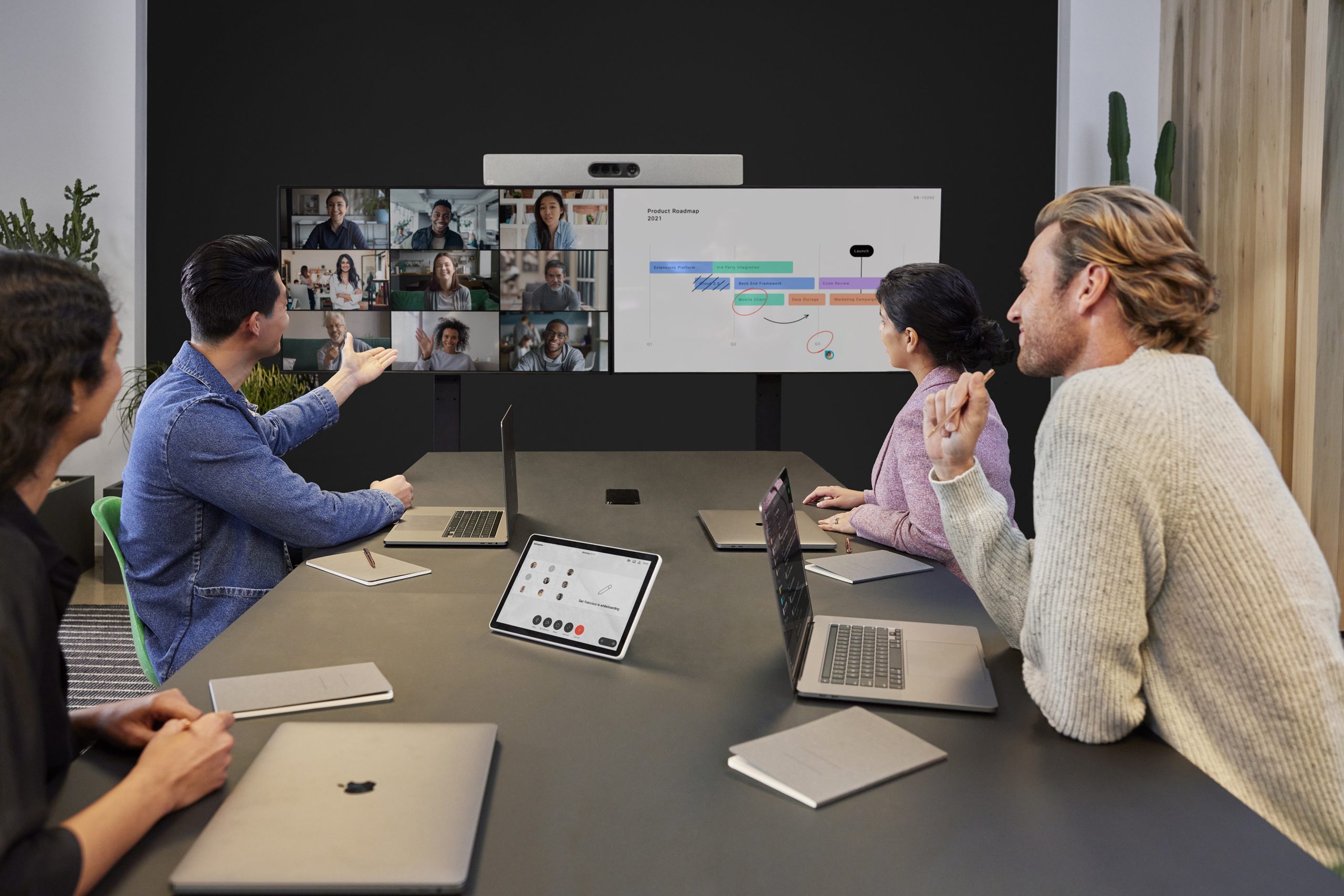图片展示了四位专业人士在办公室开会，使用双屏幕视频会议系统，讨论图表和数据，氛围看起来专注而合作。