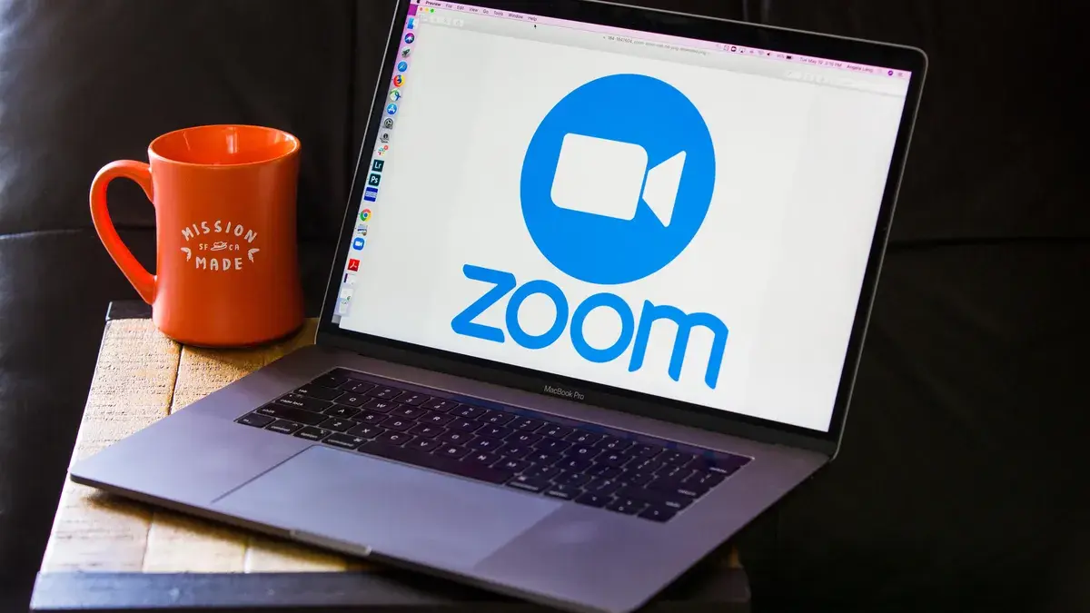 这是一台笔记本电脑屏幕显示Zoom会议软件的标志，旁边是一个橙色的咖啡杯，放在木质桌面上，背景为黑色。