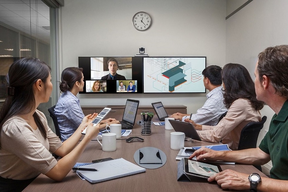 图片展示了一间会议室，五人正通过视频会议系统与远程参与者交流，墙上挂着时钟，桌上摆放笔记本和平板电脑。