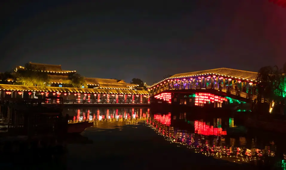 这是一张夜景照片，显示了一座装饰着五彩缤纷灯光的桥梁，其倒影在水面上显得格外迷人，周围建筑亦灯火辉煌。