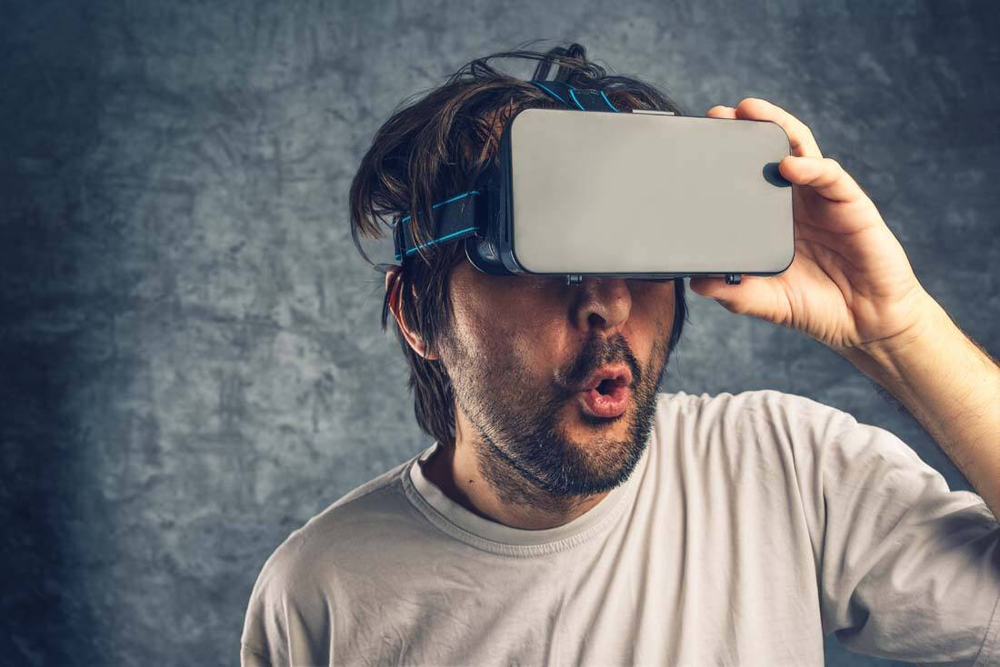 一位男士用手机遮住双眼，似乎在模拟虚拟现实体验，表情惊讶，手机前端有蓝色绑带固定在头上。