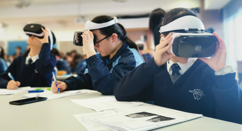 图片展示几位穿着校服的学生在教室内戴着虚拟现实头盔体验VR技术，其中一人正手持笔做记录。