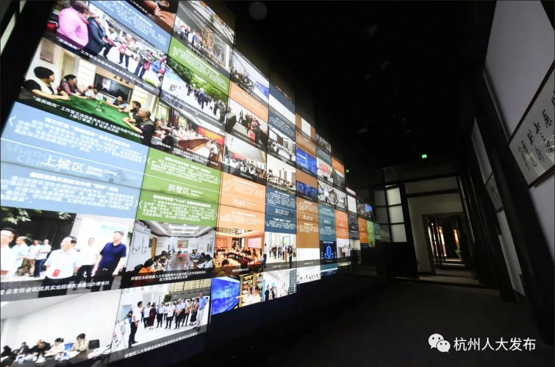 这是一面展示多个屏幕的墙，屏幕上显示各种图片和文字，内容涉及社会活动和人物，场景显得现代化和信息化。