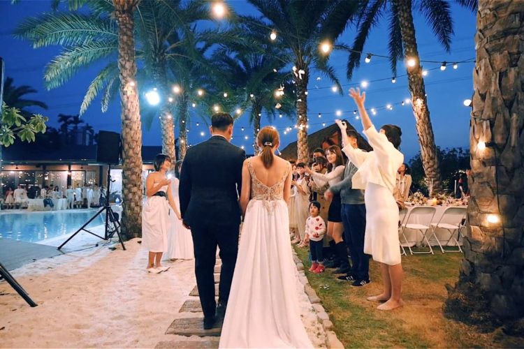 一对新人在夜晚户外举行婚礼，走过铺有沙子的通道，两旁是热情祝福的宾客和摇曳的棕榈树。