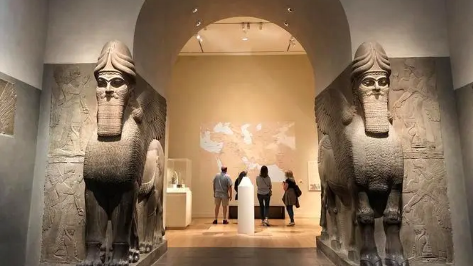图片展示了博物馆内的展厅，中间有两尊巨大的石雕像，几位参观者正在观看并了解这些展品。背景是世界地图。