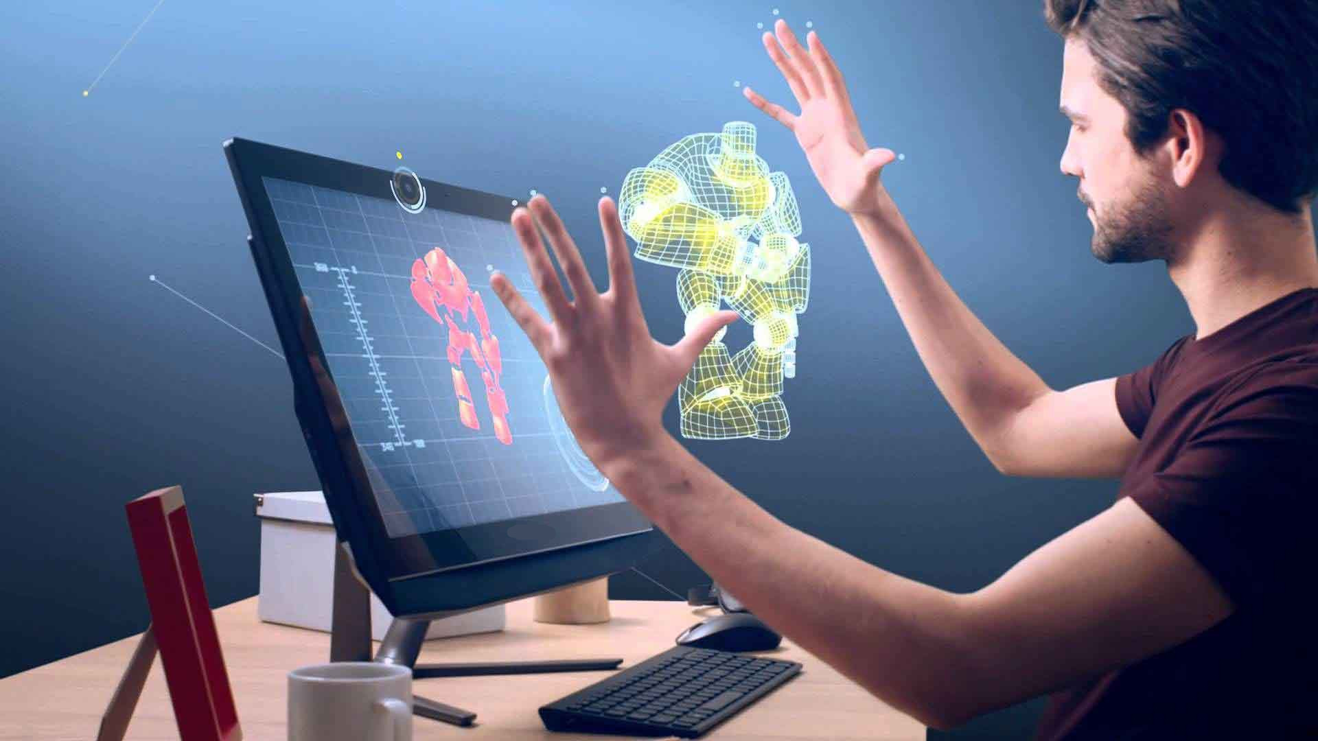 图片展示一位男士在电脑前，似乎在用手势操控屏幕上的三维图形设计。环境现代，技术感强。