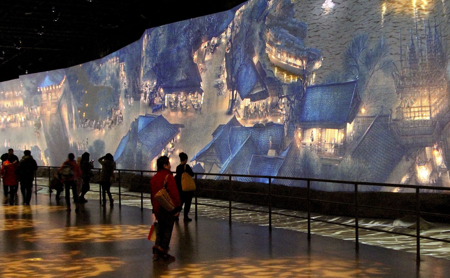 图片展示了几位参观者在观赏一幅巨大的、描绘古代山水建筑的壁画。环境昏暗，壁画细节丰富，色彩斑斓。