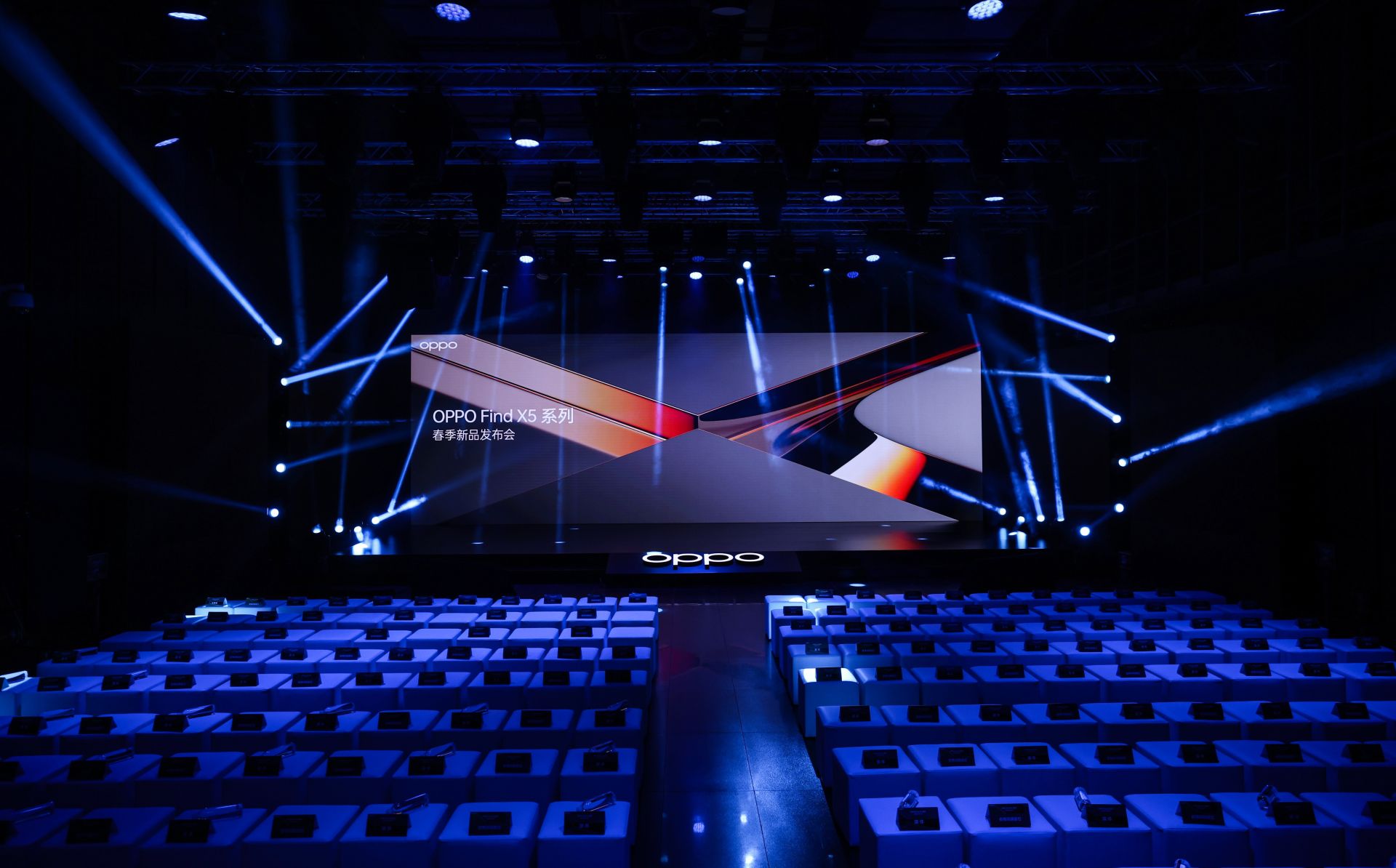 这是一场活动现场，有蓝色灯光、大屏幕显示OPPO标志，前方是整齐排列的空座位。氛围现代科技感十足。