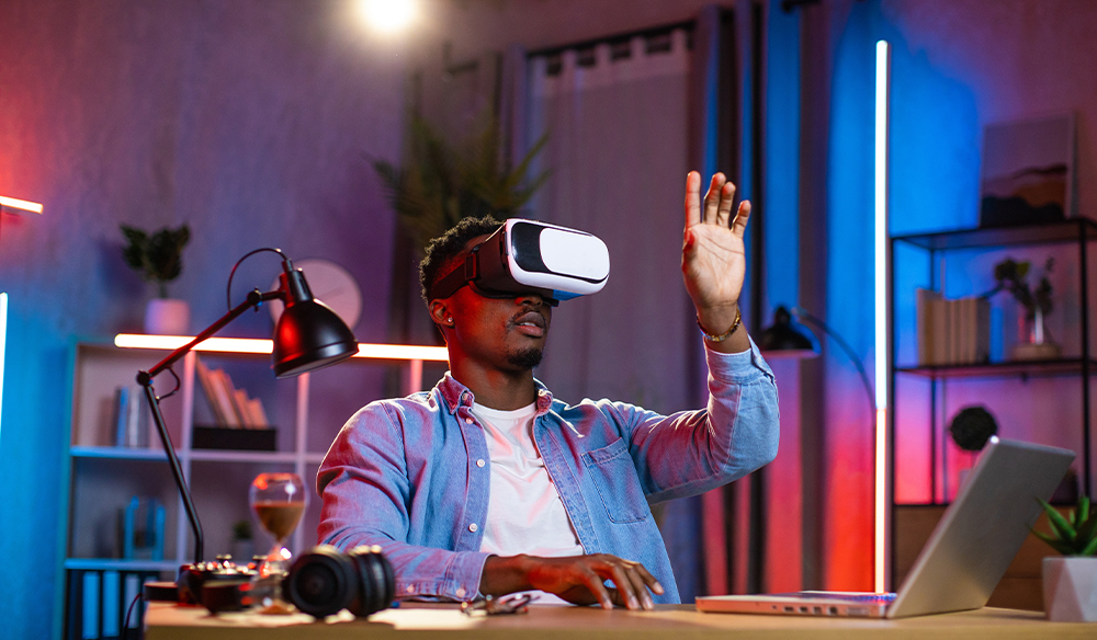 图片展示一位男士戴着虚拟现实头盔，伸手触摸虚构物体，桌上有笔记本电脑和相机，背景是彩色灯光照亮的房间。