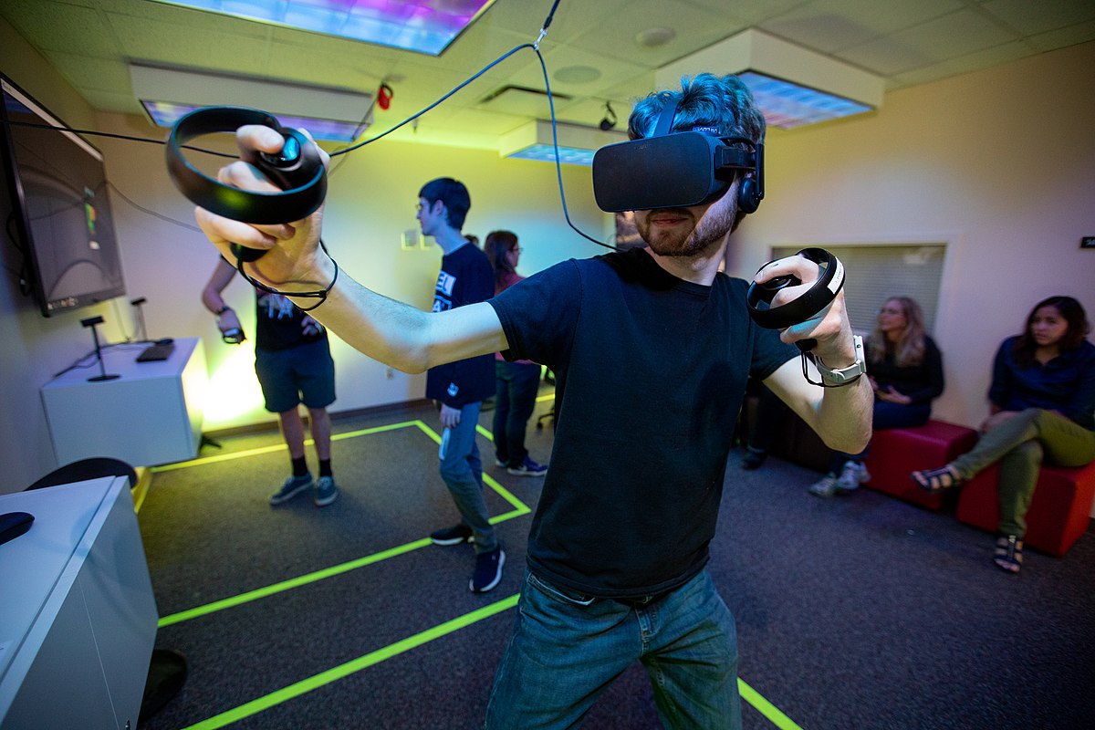 图片展示一位戴着虚拟现实头盔的人正在体验VR游戏，周围有其他人在观看。他似乎正在享受沉浸式的互动体验。