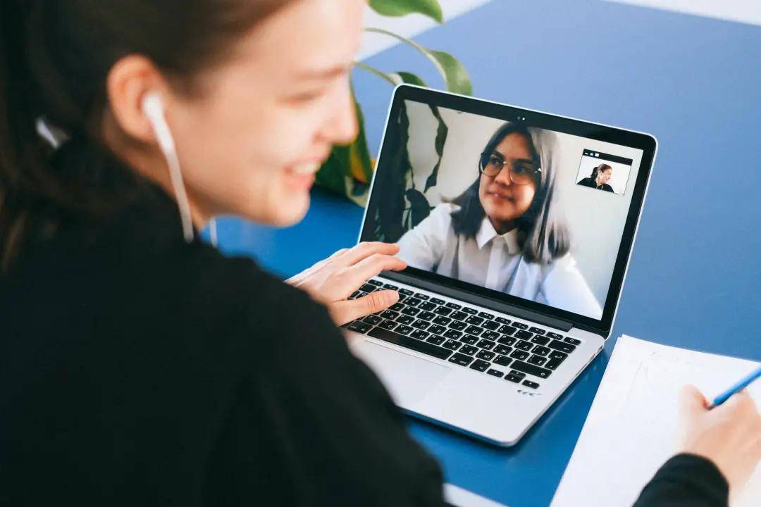 一位女士正在笔记本电脑上进行视频通话，她正佩戴耳机微笑着，电脑屏幕显示另一位女性。旁边有植物和笔记本。