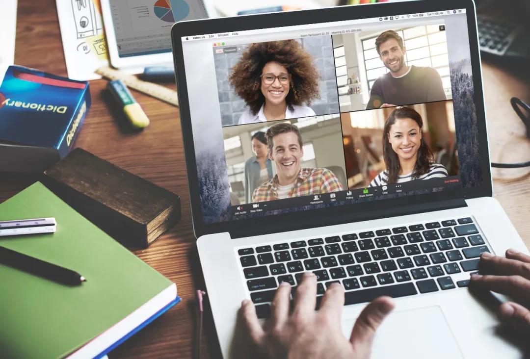笔记本电脑屏幕上显示着四个微笑的人的视频会议，桌上散落着办公用品，如笔记本和手机。