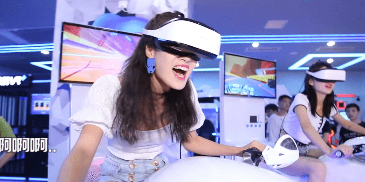 图片展示两位女性戴着VR头盔体验虚拟现实游戏，其中一位露出兴奋的表情，背景是模糊的旁观者和电子屏幕。